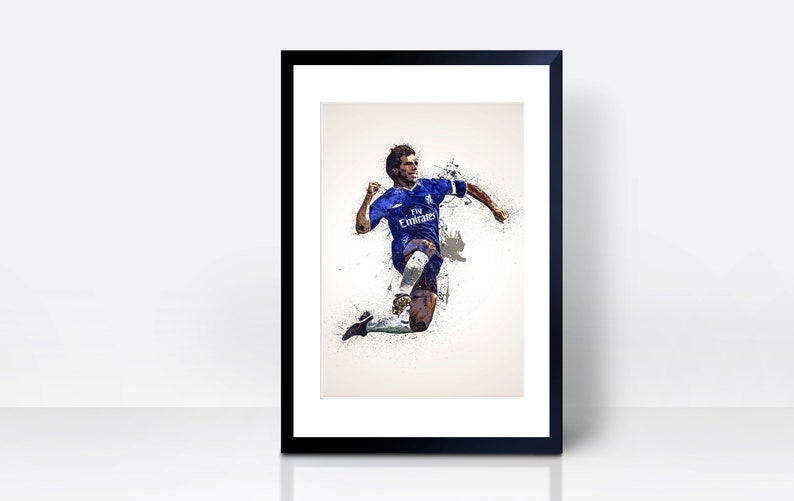 May include: A framed print of a soccer player in a blue jersey with white accents, running with the ball. The player is in mid-air, kicking the ball with his right foot. The background is a white splatter effect.