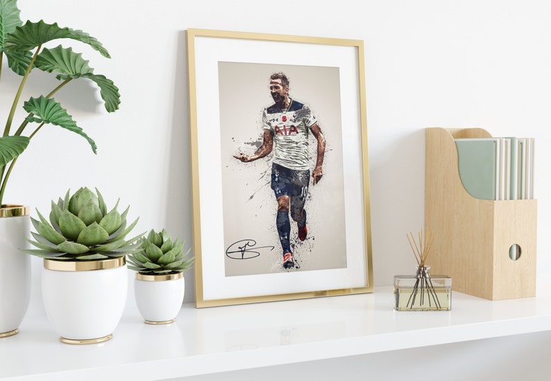 May include: A framed print of a football player in a white shirt with a blue and white pattern, running with the ball. The player's shirt has the text "AIA" on the front. The print has a gold frame and is on a white shelf with plants and a wooden box.