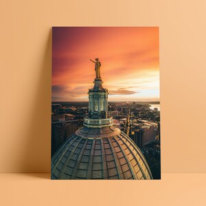 May include: An aerial view of a city skyline at sunset, with a golden statue atop a domed building. The sky is a vibrant orange and pink, with clouds reflecting the warm light.