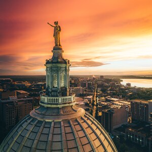 May include: An aerial view of a city skyline at sunset with a golden statue atop a large, domed building. The sky is a vibrant orange and pink, and the city lights are visible in the distance.