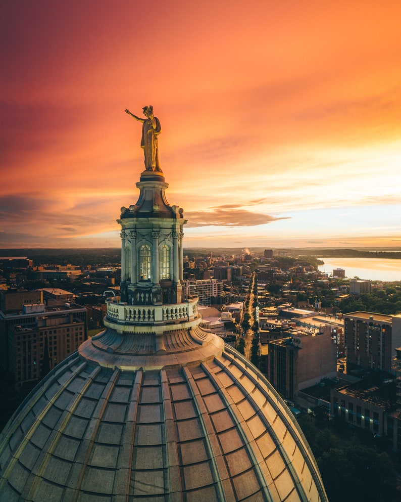 May include: An aerial view of a city skyline at sunset with a golden statue atop a large, domed building. The sky is a vibrant orange and pink, and the city lights are visible in the distance.