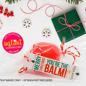 May include: A red lip balm wrapped in clear cellophane with a white tag that reads "Merry Christmas You're the Balm!" with a small Christmas tree graphic. The tag is tied with red and white twine. The lip balm is a Christmas gift with a personalized tag.