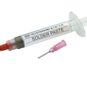 Könnte beinhalten: Eine transparente Plastikspritze gefüllt mit grauer Lötpaste, beschriftet mit "HILDERBRAND & CIE S.A. SOLDER PASTE". Eine separate rosa Plastiknadelspitze ist im Lieferumfang enthalten.