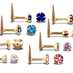 May include: An assortment of gold-coloured earrings featuring studs with gemstones and a heart shape. Gemstones are in blue, red, pink, and purple. The earrings are presented against a white backdrop.