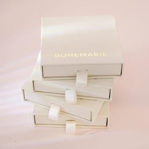 May include: A stack of four square, off-white jewelry boxes with sliding drawers. Each box features a small, matching off-white fabric pull tab. The top box displays the brand name BOHEMARIE printed in gold lettering on the front surface.