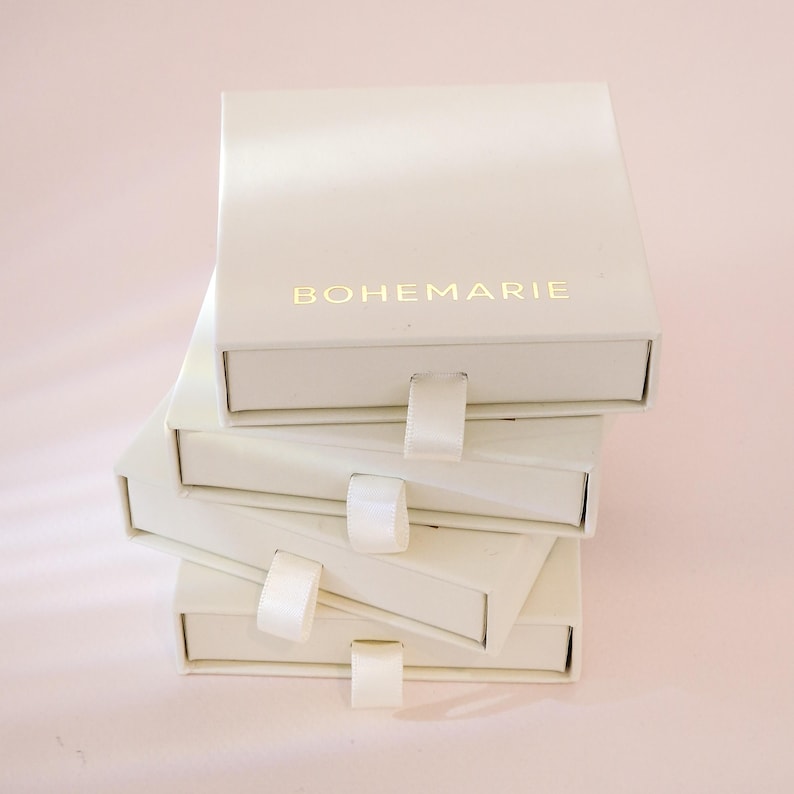May include: A stack of four square, off-white jewelry boxes with sliding drawers. Each box features a small, matching off-white fabric pull tab. The top box displays the brand name BOHEMARIE printed in gold lettering on the front surface.