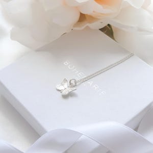 May include: A silver butterfly pendant necklace on a white box with the text "BOHEMARIE". The butterfly pendant is detailed with etched wings and hangs from a delicate chain. A white ribbon and a flower are in the background.
