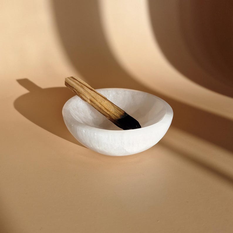 May include: A white, round, carved stone bowl with a Palo Santo stick. The Palo Santo stick is brown with a black end. The bowl is on a tan surface with shadows.