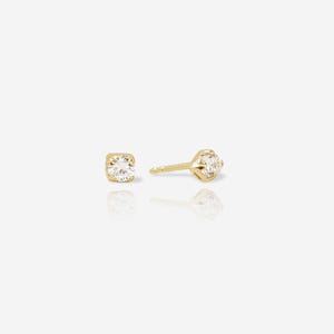 May include: A pair of gold stud earrings, each featuring a single round, clear, faceted gemstone set in a four-prong gold setting. The earrings are shown from the front and side, with a post and butterfly back visible on the side view.