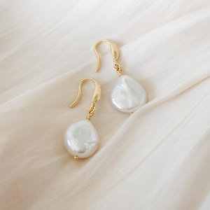 May include: A pair of gold-colored drop earrings with large, irregular white pearls. The earrings are displayed on a cream-colored fabric, showcasing their elegant design and natural pearl texture. The hooks are curved.
