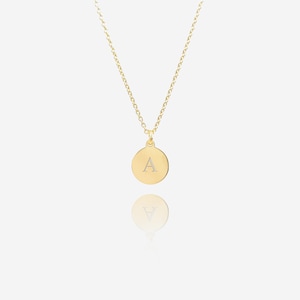 May include: A gold-coloured necklace with a circular pendant. The pendant displays the letter "A" in a lighter shade. The necklace chain is fine and suspends the pendant. The background is light, and the pendant's reflection is visible.