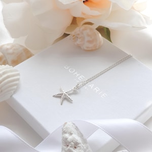 May include: A silver starfish pendant necklace on a white box with the word "Bohemarie" printed on it. The necklace is displayed with white flowers, seashells, and a white ribbon.