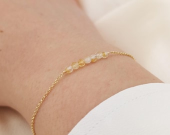 May include: A delicate gold bracelet featuring small, round, yellow and clear gemstones. The bracelet is strung on a fine gold chain, creating a minimalist and elegant design. The gemstones are evenly spaced along the chain.