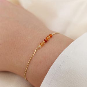 May include: A delicate gold chain bracelet featuring a series of small, round, orange and yellow gemstone beads. The bracelet is displayed on a wrist, showcasing the jewelry's design and color.