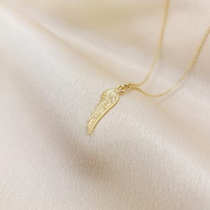 May include: A gold-colored necklace featuring a detailed wing pendant. The wing design has intricate feather detailing and hangs from a delicate chain. The necklace is displayed on a soft, cream-colored fabric.