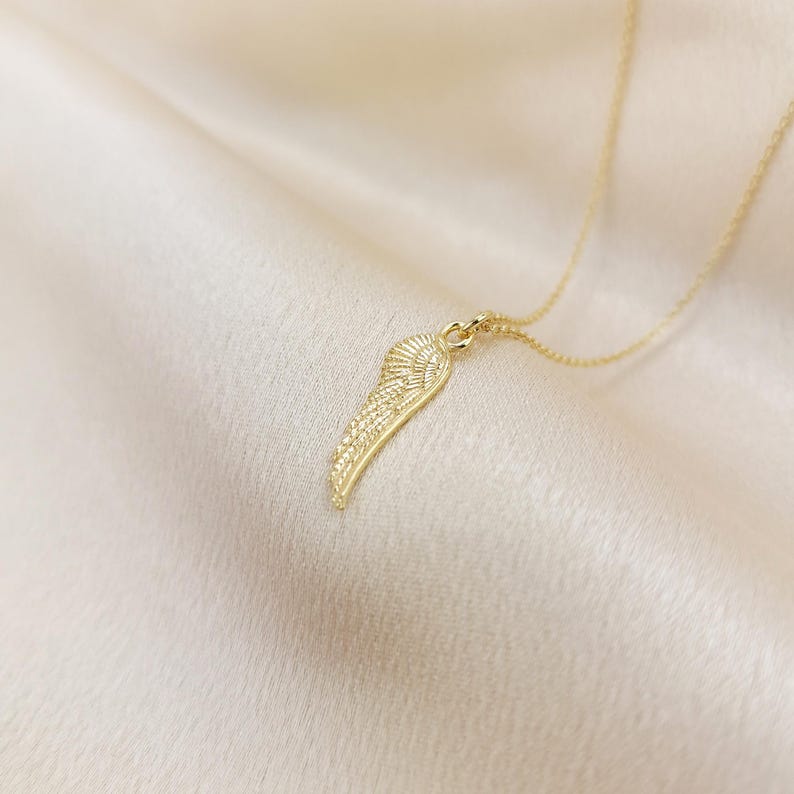 May include: A gold-colored necklace featuring a detailed wing pendant. The wing design has intricate feather detailing and hangs from a delicate chain. The necklace is displayed on a soft, cream-colored fabric.