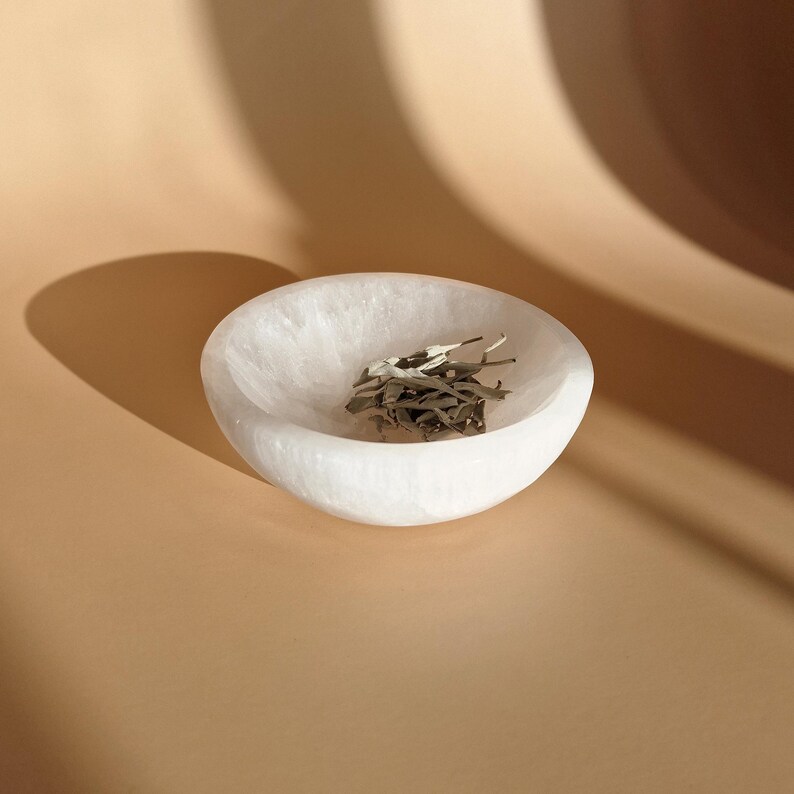 May include: A white, oval-shaped stone bowl filled with dried green leaves. The bowl has a smooth, polished interior and sits on a light brown surface. The lighting creates a shadow on the background.