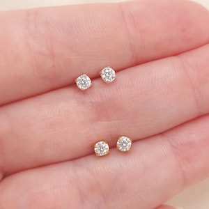 May include: Two pairs of stud earrings featuring round, clear, faceted stones set in four-prong metal settings. One pair is set in silver-toned metal, and the other pair is set in gold-toned metal, both displaying a brilliant, reflective surface.