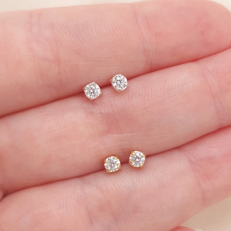 May include: Two pairs of stud earrings featuring round, clear, faceted stones set in four-prong metal settings. One pair is set in silver-toned metal, and the other pair is set in gold-toned metal, both displaying a brilliant, reflective surface.