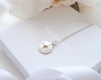 May include: A silver necklace featuring a circular pendant with a cut-out star shape in the center and the letters N, E, S, and W engraved around the edges. The pendant rests on a white box with the text BOHEMARIE printed on the top surface.
