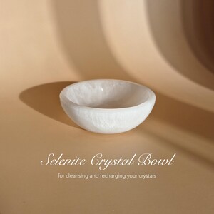 May include: A white, translucent selenite crystal bowl with a smooth, rounded interior and exterior. The text "Selenite Crystal Bowl" and "for cleansing and recharging your crystals" is printed in white below the bowl.