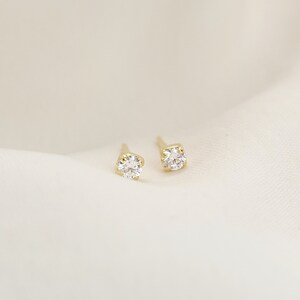 May include: A pair of gold stud earrings, each featuring a single round, clear, faceted gemstone held in a four-prong setting. The earrings are positioned side by side, showcasing the metallic yellow gold posts and the brilliant cut of the stones.