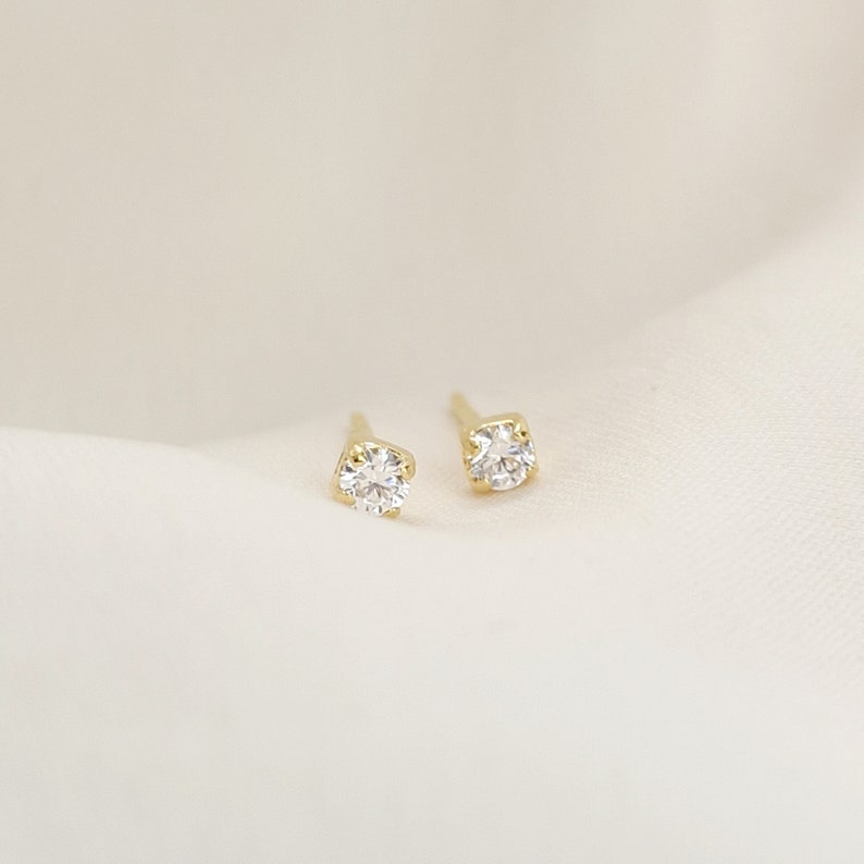 May include: A pair of gold stud earrings, each featuring a single round, clear, faceted gemstone held in a four-prong setting. The earrings are positioned side by side, showcasing the metallic yellow gold posts and the brilliant cut of the stones.