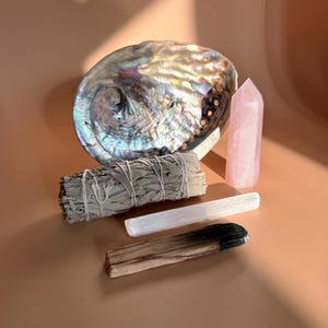 May include: An arrangement of spiritual items on a peach-colored surface. Includes a large iridescent abalone shell, a bundle of sage, a rose quartz crystal, a selenite wand, and a palo santo stick.