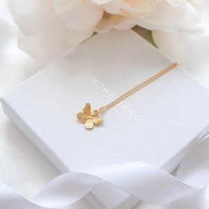 May include: A gold-toned necklace featuring a butterfly pendant with detailed wing textures. The necklace rests on a white square box with the text BOHEMARIE printed on the top surface. The jewelry piece is delicate with a thin chain link design.