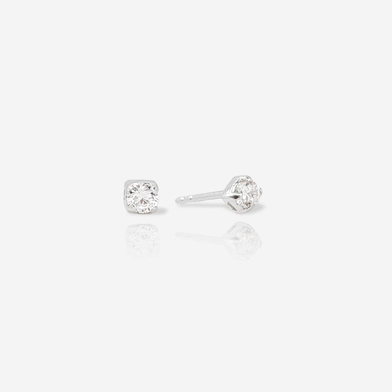 May include: A pair of silver stud earrings featuring a single round, clear, faceted gemstone set in a four-prong metal setting. One earring is shown from the front, while the other is positioned to display the post and the side profile of the setting.