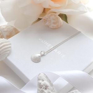 May include: A silver necklace featuring a small, textured seashell pendant. The necklace rests on a white square jewelry box with the text BOHEMARIE printed in silver. The design includes a delicate chain and a realistic, ridged shell charm.