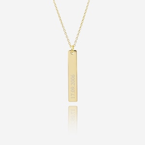 May include: A gold-coloured vertical bar pendant necklace with a delicate chain. The pendant has the engraved date "17.09.2006". The necklace is displayed on a white background with a reflection.