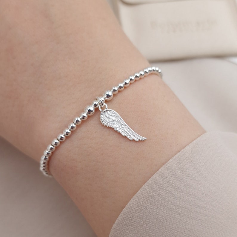 May include: A silver beaded bracelet with a silver angel wing charm. The bracelet is on a person's wrist.