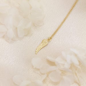 May include: A gold-colored necklace featuring a delicate wing pendant. The wing is intricately detailed, with a textured surface. The necklace hangs on a fine chain, and the background is a soft, off-white fabric. The image is well-lit, highlighting the jewelry's design.