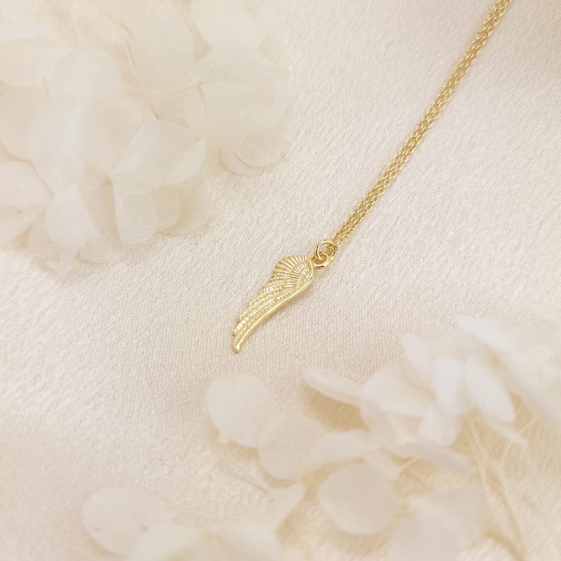 May include: A gold-colored necklace featuring a delicate wing pendant. The wing is intricately detailed, with a textured surface. The necklace hangs on a fine chain, and the background is a soft, off-white fabric. The image is well-lit, highlighting the jewelry's design.
