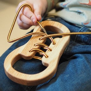 Könnte beinhalten: Ein hölzerner Spielzeugschuh mit gelben und braunen Schnürsenkeln. Eine Kinderhand fädelt die Schnürsenkel durch den Schuh.