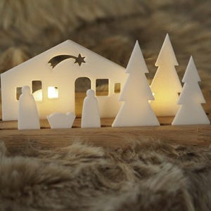Könnte beinhalten: Weiße Weihnachtskrippe aus Holz mit einem Stern, einem Haus, drei Bäumen und Figuren von Maria, Josef und dem Jesuskind. Die Szene ist von innen beleuchtet und erzeugt ein warmes Leuchten.