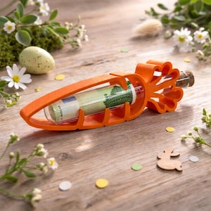Könnte beinhalten: Ein orangefarbener, karottenförmiger Geldhalter mit einem Glasröhrchen im Inneren, das eine aufgerollte Banknote enthält. Der Halter hat an einem Ende einen Korken und steht auf einer Holzoberfläche mit Osterdekorationen.