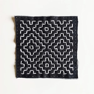 May include: A square, black fabric patch with a white geometric embroidered design. The design features repeating stepped patterns and square shapes, creating a symmetrical pattern. The edges of the patch are finished with a simple stitch.