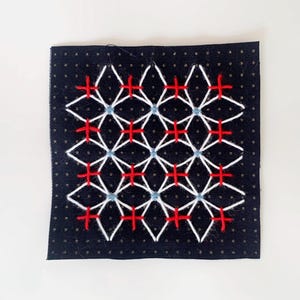 Könnte beinhalten: Ein schwarzes Stoffquadrat mit einem geometrischen Muster, das mit weißem, rotem und blauem Garn gestickt ist.