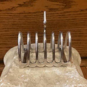 May include: A silver-colored metal toast rack with six slots, set on a light-colored, scalloped base. The rack has a handle and is designed to hold slices of toast. The background is a wooden surface.