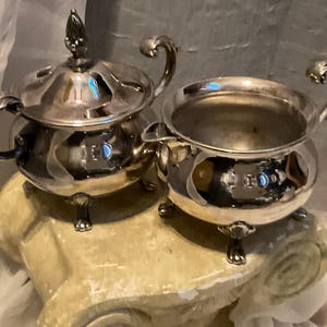 May include: A silver tea set, including a covered teapot and a creamer, sits on a decorative stand. The teapot has a decorative finial and curved handle. The creamer has a curved handle. Both pieces have small feet.