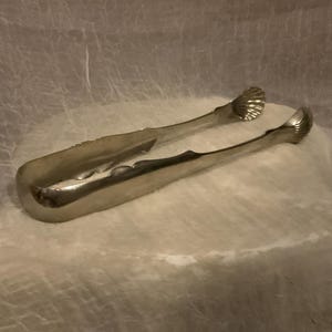 May include: Silver-toned sugar tongs with a scalloped design on the gripping ends. The tongs are curved and have a decorative edge along the handle. The image is set against a soft, textured background.