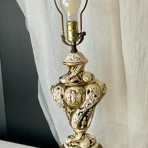 May include: A table lamp featuring a cream-colored ceramic base with intricate cut-out details, gold accents, and a central medallion depicting two figures. The lamp includes a brass-toned metal harp, a light bulb, and a hexagonal metal base.