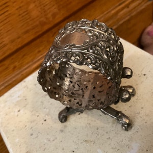 May include: Silver napkin ring with an ornate design and three legs. The ring is made of a delicate, filigree metal and has a slightly tarnished finish.