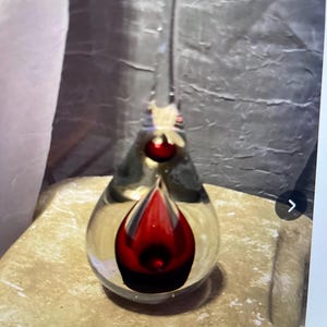 May include: A clear glass art piece with a teardrop shape, featuring a vibrant red interior design. The piece has a long, slender top and a rounded base. The glass is transparent, allowing the red design to be visible. The art piece is displayed on a stone surface.
