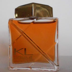 Peut inclure: Un flacon de parfum carré avec un bouchon doré et un corps en verre transparent. Le flacon contient un liquide de couleur orange. Le texte "KL Homme" est imprimé sur le devant du flacon.