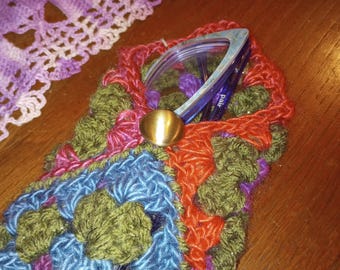 May include: A crocheted glasses case with a colorful geometric pattern in shades of green, blue, orange, and purple. The case has a metal clasp and is holding a pair of blue glasses.