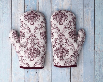 Puede incluir: Un par de guantes para horno con un patrón floral burdeos sobre un fondo beige claro. Los guantes tienen un ribete burdeos.