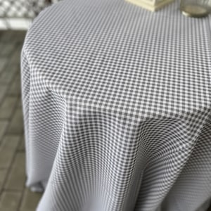 Puede incluir: Un mantel blanco con un patrón de cuadros grises. El mantel está drapeado sobre una mesa redonda.